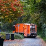 orange trash truck parks on a quiet suburban neighborhood street. Trash cans in the foreground have just been emptied.