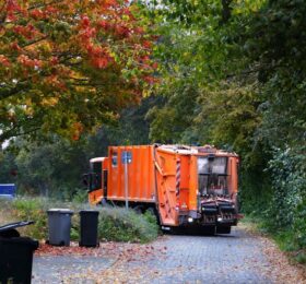 orange trash truck parks on a quiet suburban neighborhood street. Trash cans in the foreground have just been emptied.
