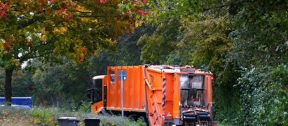 orange trash truck parks on a quiet suburban neighborhood street. Trash cans in the foreground have just been emptied.