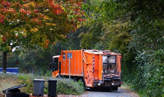 orange trash truck parks on a quiet suburban neighborhood street. Trash cans in the foreground have just been emptied.